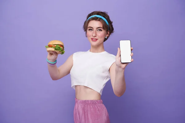 Happy woman holding hamburger and showing food delivery app on mobile phone to order lunch isolated on studio background.