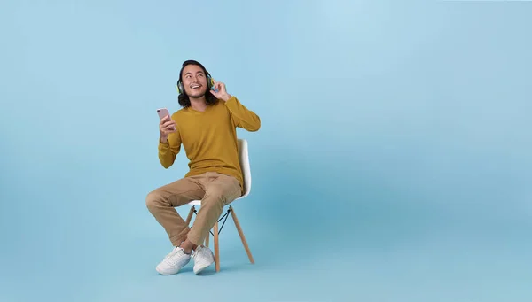 Joyful young Asian guy in wireless headphones with mobile phone sitting on chair, having fun smile, listening to music, enjoying cool soundtrack against on blue studio empty space