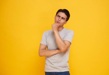Düşünceli, karmaşık Asyalı bir adam, çenesine dokunuyor ve sol üst köşeye bakıyor, düşünüyor, karar veriyor, bir şeyler seçiyor, şüpheleri var, sarı arka planda tereddütlü duruyor..