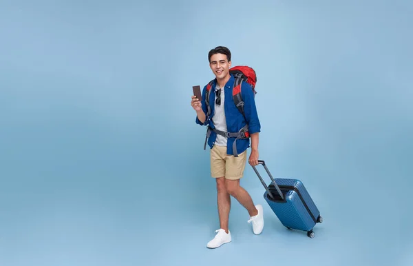 完全な長さ幸せな笑顔の若い観光客アジアの男は荷物を保持しながら歩くと孤立したスタジオ青の背景にパスポートを示す — ストック写真