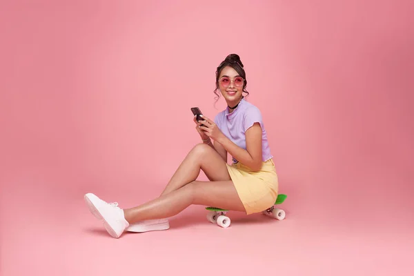 スマートフォンを使用し パステルピンクの背景に隔離されたスケートボードに座っているフル長さ幸せな若いアジアの女性の女の子 人々のライフスタイルの概念 スケートボードに座る — ストック写真