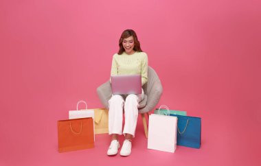 Güzel Asyalı genç kadın kanepeye oturmuş pembe arka planda alışveriş torbalarıyla internetten laptop alışverişi yapıyor..