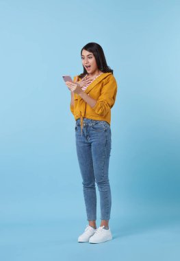 Mutlu bir Asyalı portresi güzel bir genç kız gülümsüyor. Şaşırmış bir şekilde mavi stüdyo arka planında izole edilmiş akıllı cep telefonu kullanıyor..