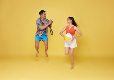 Mutlu Asyalı çiftler seyahat etmek için zıplıyor ve sarı arka planda izole bir yaz tatilinde fotoğraf çekiyor..