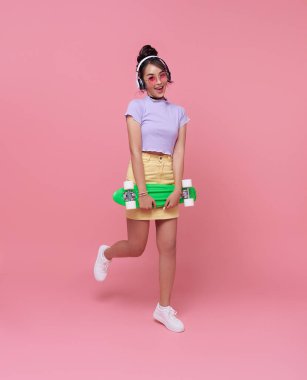 Genç Asyalı genç kız elinde kaykayla kulaklık takmış pembe arka planda müzik dinliyor. 20 'li yaşlarda kadın