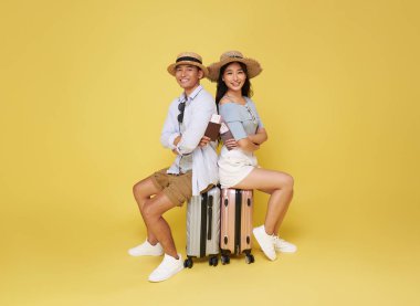 Mutlu gülümseyen genç Asyalı çift bavulları üzerinde oturuyor ve ellerinde pasaportlarıyla seyahate çıkıyorlar sarı stüdyo arka planında izole bir şekilde..