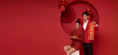 Kırmızı günlük kıyafetler içinde mutlu Asyalı çift angpao ya da kırmızı paket para hediyesi taşıyorlar ve altın külçeler mesajla kutsanmış bu da kırmızı oda dekorasyonunda Çin 'in yeni yılı için büyük şans demek..