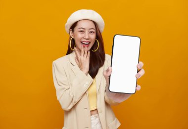 Güzel Asyalı iş kadını elinde akıllı telefon ile boş ekranın bir kopyasını tutuyor ve sarı arka planda gülümsüyor..