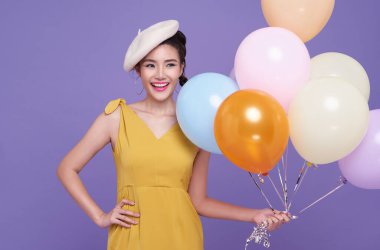 Gülümseyen mutlu, mutlu, sarı elbiseli Asyalı kadın mor arka planda izole edilmiş balonları kutluyor. Doğum günü ve yeni yıl partisi konsepti