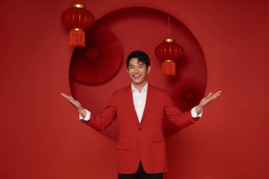Yeni Çin yılın kutlu olsun. 2024 yılının Çin yılbaşı jesti ile kırmızı takım elbise giyen Asyalı adam kırmızı dekorasyon arka planında izole edilmiş..