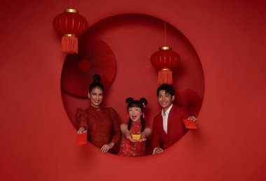 Kırmızı zarf taşıyan geleneksel Cheongsam Qipao elbisesi giyen mutlu Asyalı bir aile. ang pao, Çin 'in 2024 yılındaki yeni yılı için kırmızı dekorasyon arka planında izole edilmiş kırmızı bir zarf verdi..