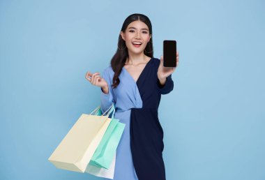 Neşeli, mutlu Asyalı alışveriş yapan kadın alışveriş torbaları taşıyor ve mavi arka planda izole akıllı telefon ekranını gösteriyor..