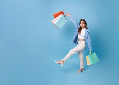 Uzun boylu, Asyalı alışverişçi kadın elinde alışveriş çantası ile geziyor. Mavi fotokopi arkaplanından izole edilmiş indirimli promosyon için heyecanlı..