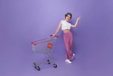 Alışveriş arabasının işaret parmağı mor arka planda izole edilmiş alışveriş merkezinde tam boy, neşeli bir kadın..