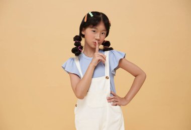 Asyalı küçük kız parmaklarını dudaklarının üzerinde tutuyor, sessiz jestler yapıyor, dedikodular çıplak renk arka planında izole edilmiş..