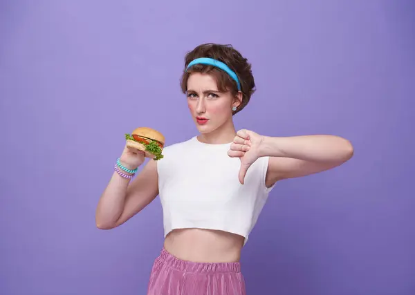 Young sad woman hold eat burger show hand stop gesture say no isolated on purple background. Proper nutrition healthy lifestyle fast food unhealthy choice concept