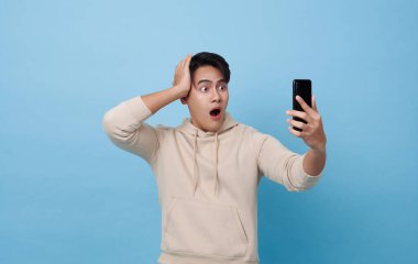 Asyalı adamın yüzü mavi arka planda izole akıllı telefonda gördüklerini şok etti..