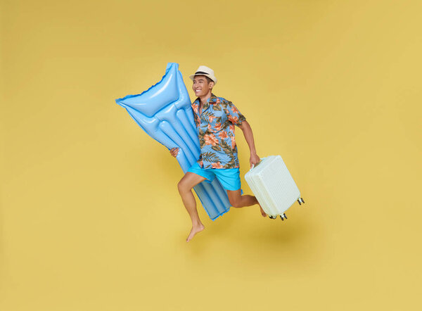 Full body young traveler tourist asian man in summer clothes hat hold suitcase inflatable mattress run isolated on yellow background. 