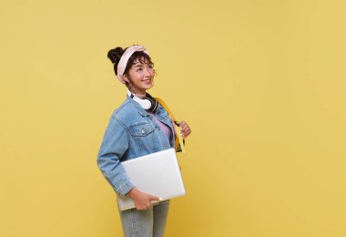 Sırt çantası ve dizüstü bilgisayarı olan mutlu gülümseyen Asyalı üniversite öğrencisinin portresi sarı arka planda izole bir şekilde okula hazır..