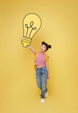 Happy Asian child student holding light bulb isolated on yellow background. idea thinking and creative concept.