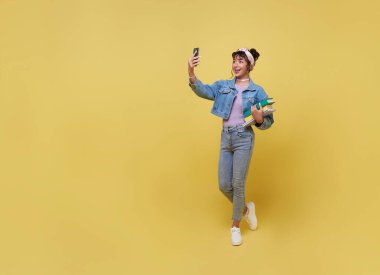 Portrait of happy smiling young Asian college student holding books and  taking selfie with smartphone isolated on yellow background.