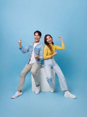 Happy Asian couple sitting back to back on white cube chair doing winner gesture celebrate clenching fists say yes isolated on blue studio background.