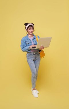 Sarı arka planda, okula gitmeye hazır dizüstü bilgisayar ve sırt çantası kullanan mutlu gülümseyen Asyalı üniversite öğrencisinin portresi. Okula geri dön.