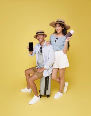 Mutlu Asyalı çift, sarı stüdyo arka planında izole edilmiş seyahat için hazır akıllı telefon modelini gösteren bavullarıyla seyahate çıkıyor..