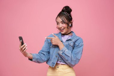 Genç Asyalı kadın akıllı telefonu işaret ederek sosyal medyayı kontrol ediyor pembe arka planda mutlu bir gülümseme var. Mutlu, sevimli, mutlu kadın başarıya sevinir..