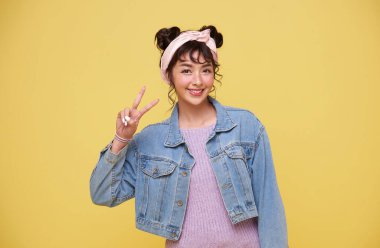 Genç, Asyalı, sıradan kıyafetler giymiş, gülümseyen, parmaklarını kameraya gösteren, sarı arka planda zafer işareti yapan hoş bir kadın..