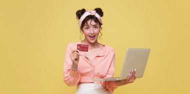 Happy beautiful Asian teen shopper women showing credit card payment for shopping online with laptop computer isolated on yellow background. clipart