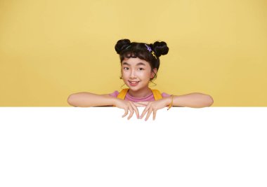 Smiling happy cute Asian student elementary school girl hiding behind a blank white board isolated on yellow background.