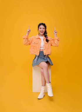 Sandalyeye oturup elini uzatarak sarı arka planda izole edilmiş mutlu Asyalı genç kız. Halk yaşam tarzı konsepti.