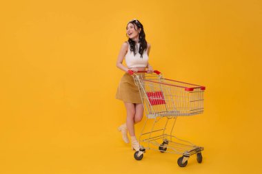 Uzun, mutlu Asyalı kadın boş alışveriş arabasını iterken ya da alışveriş arabasında yürürken sarı arka planda izole edilmiş süpermarkette..