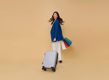 Mutlu, genç, zarif Asyalı iş kadını seyahat valizleri ve alışveriş çantası çıplak renk arka planında izole edilmiş. Turist kız neşeli bir tatil konsepti yaşıyor. 