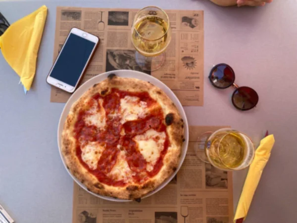 Blurred background of a table in a pizzeria with pizza, mobile phone and sunglasses, save space. Vacation concept by the sea in southern country