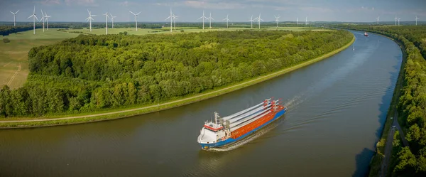 Cargo ship carrying wind turbine blades is navigating through the Kiel Canal with a wind farm in the background. Transportation of blades for wind turbines on a cargo ship.