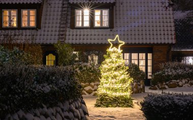 Cozy house with snow-covered roof and warmly glowing windows features decorated Christmas tree with bright lights, star on top in the front yard, surrounded by snow-dusted bushes and wintry landscape. clipart