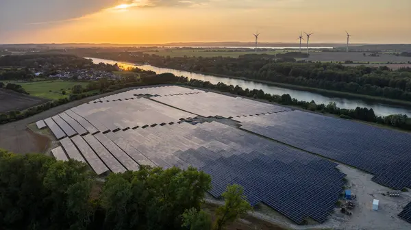 stock image Aerial view of a large solar farm with wind turbines in the background, set against a scenic landscape on canal at sunset. Ground-mounted solar photovoltaic panels neighboring canal.