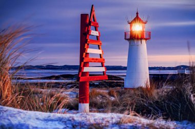 Coastal lighthouse glowing warmly against winter twilight, with striking red and white triangular navigational marker beside it, set amidst snow-dusted dunes, frosty grasses and purple skies.Sylt. clipart