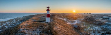 Red and white striped lighthouse situated on a coastal landscape at sunrise in winter. Lighthouse West-List on the Ellenbogen (the 