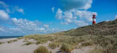 Sahil manzarasında bulunan kırmızı ve beyaz çizgili deniz feneri. Deniz feneri mavi gökyüzünün altında, geniş denize bakan çimenli kumulların üzerinde duruyor. Ellenbogen 'da Deniz Feneri Batı Listesi (