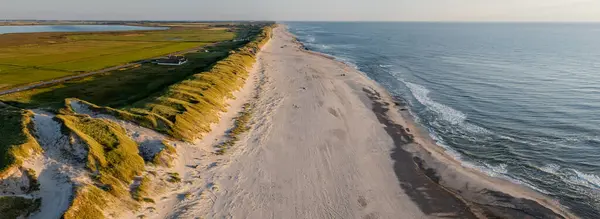 Aerial view of long, untouched coastline with lush green meadows, golden dune and sandy beach meeting sea with gentle waves on one side while tranquil waters of Ringkbing Fjord stretch on other side