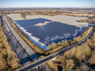 Kış manzarasında karayolu kavşağı yakınında büyük ölçekli güneş çiftliği kurulumu. Fotovoltaik panellerle güneş çiftliğinin görüntüsü kısmen, devam eden ince bir buz tabakasıyla kaplı..