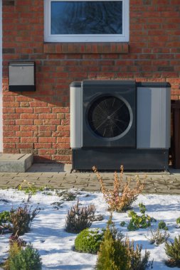 Modern heat pump unit outside brick house in winter. Heat pump unit is installed against the brick wall of a house. Modern heating and cooling technology. clipart