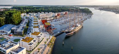 Lüks rıhtım daireleri, yelkenli yatları ve günbatımında tarihi bir gemi olan modern marina manzarası. Priwall yarımadasının turizm, emlak, seyahat, deniz manzarası