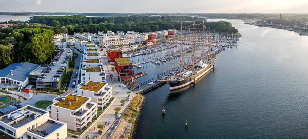 Lüks rıhtım daireleri, yelkenli yatları ve günbatımında tarihi bir gemi olan modern marina manzarası. Priwall yarımadasının turizm, emlak, seyahat, deniz manzarası