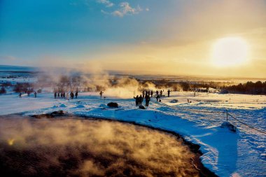 Jeotermal kaynaklı kış manzarası, turistler ve karlı sahnenin üzerine sıcak bir parıltı saçan batan güneş. Konum: Strokkur Gayzer, İzlanda.