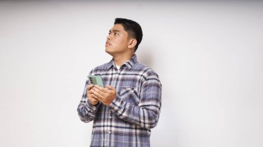 Beyaz arka planda üzgün ve kafası karışmış bir ifadeyle cep telefonunu tutan genç bir Asyalı. Stüdyo çekimi