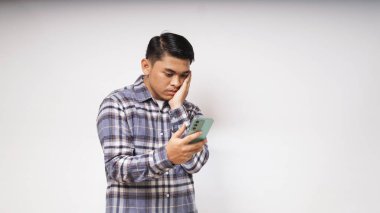 Beyaz arka planda üzgün ve kafası karışmış bir ifadeyle cep telefonunu tutan genç bir Asyalı. Stüdyo sho